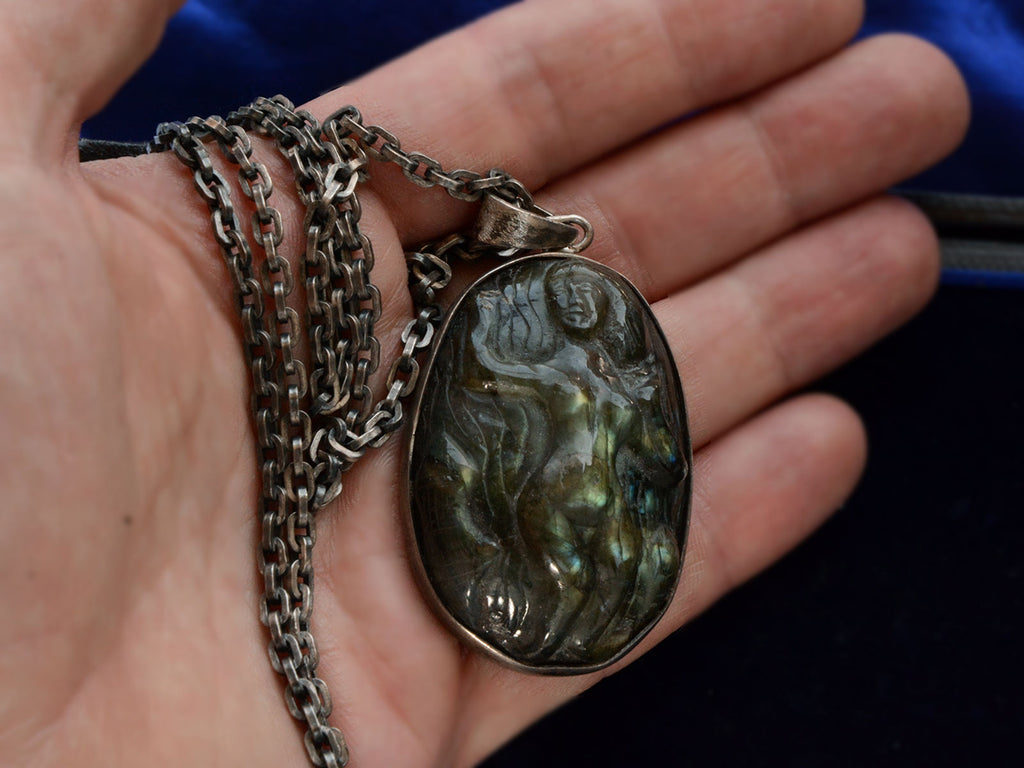 A vintage pendant necklace with labradorite carved with the image of a nude woman, shown on a hand for scale.