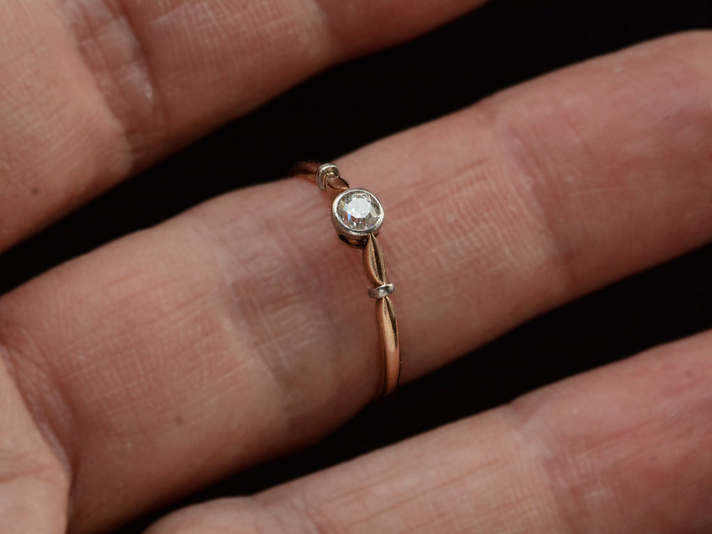 An antique Art Deco era yellow and white gold engagement ring with a bezel-set round 0.20ct old European cut diamond. Shown on finger.