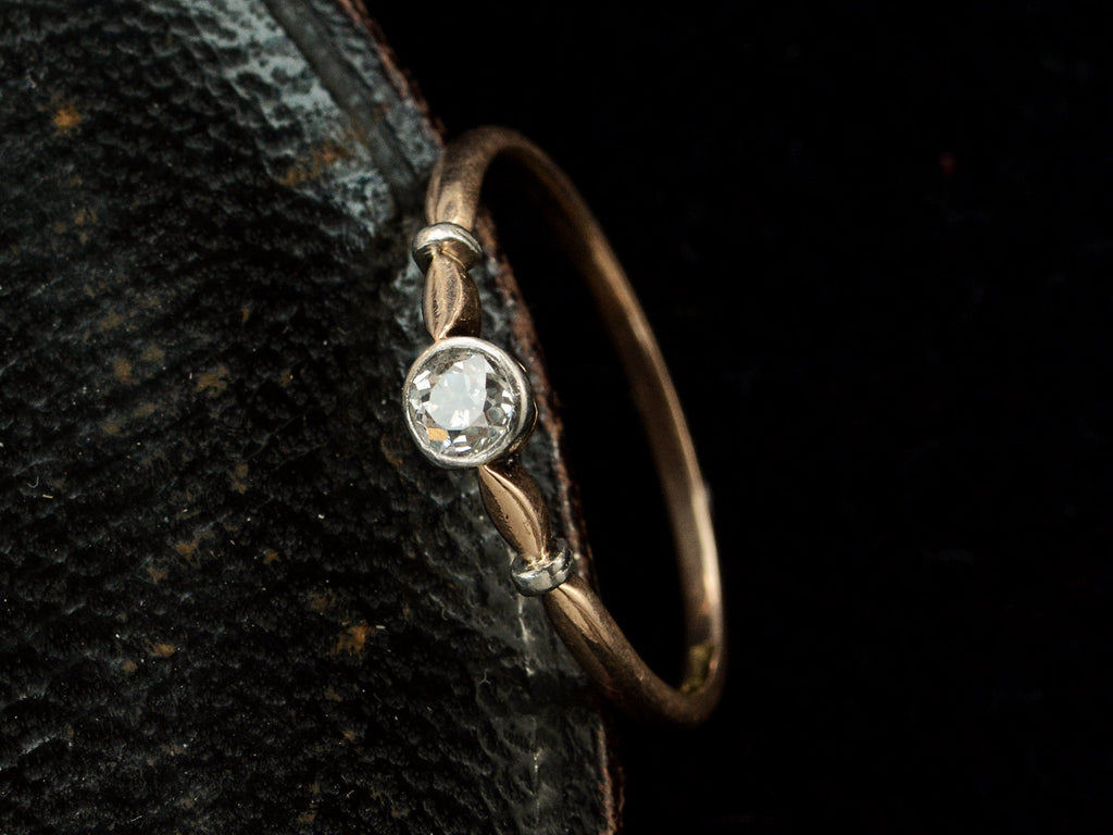 Angled view of an antique Art Deco era yellow and white gold engagement ring with a bezel-set round 0.20ct old European cut diamond. Shown on dark background.