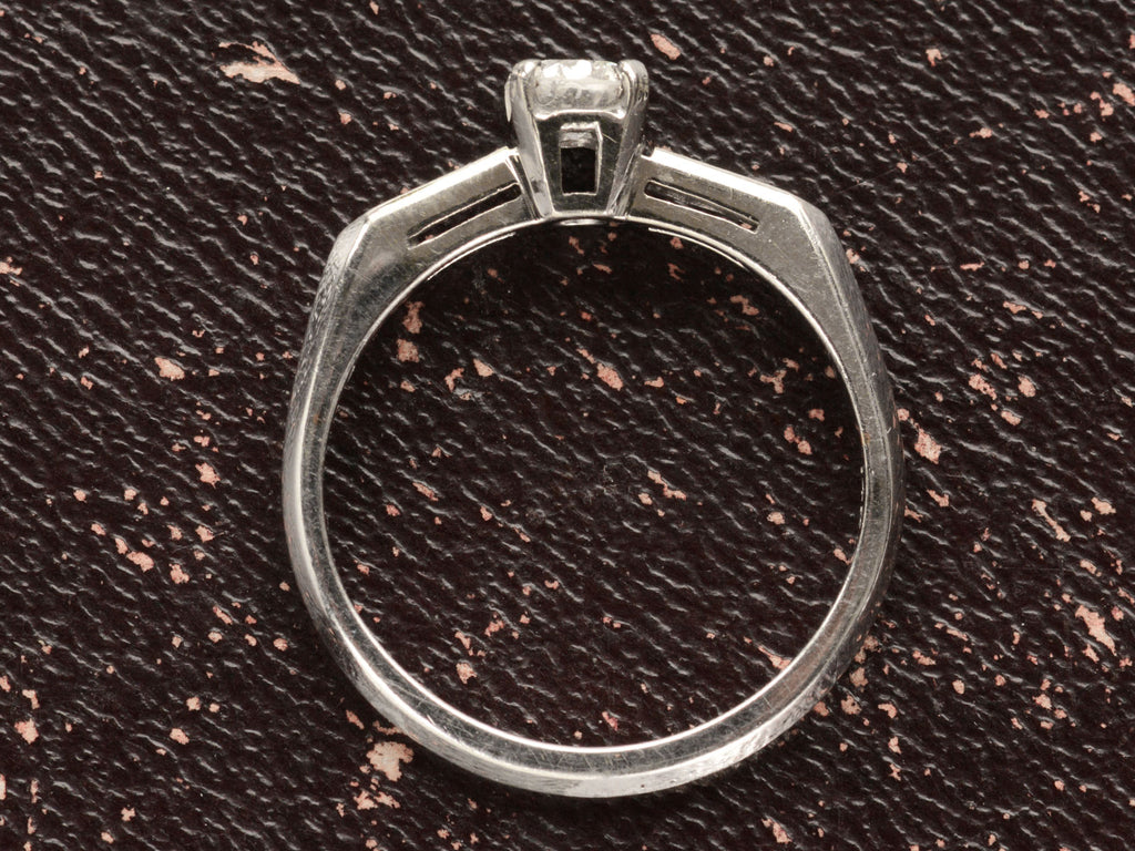 Side profile of a vintage c1940 white gold engagement ring with small round diamond and rectangular baguettes on either side. Shown on dark background.