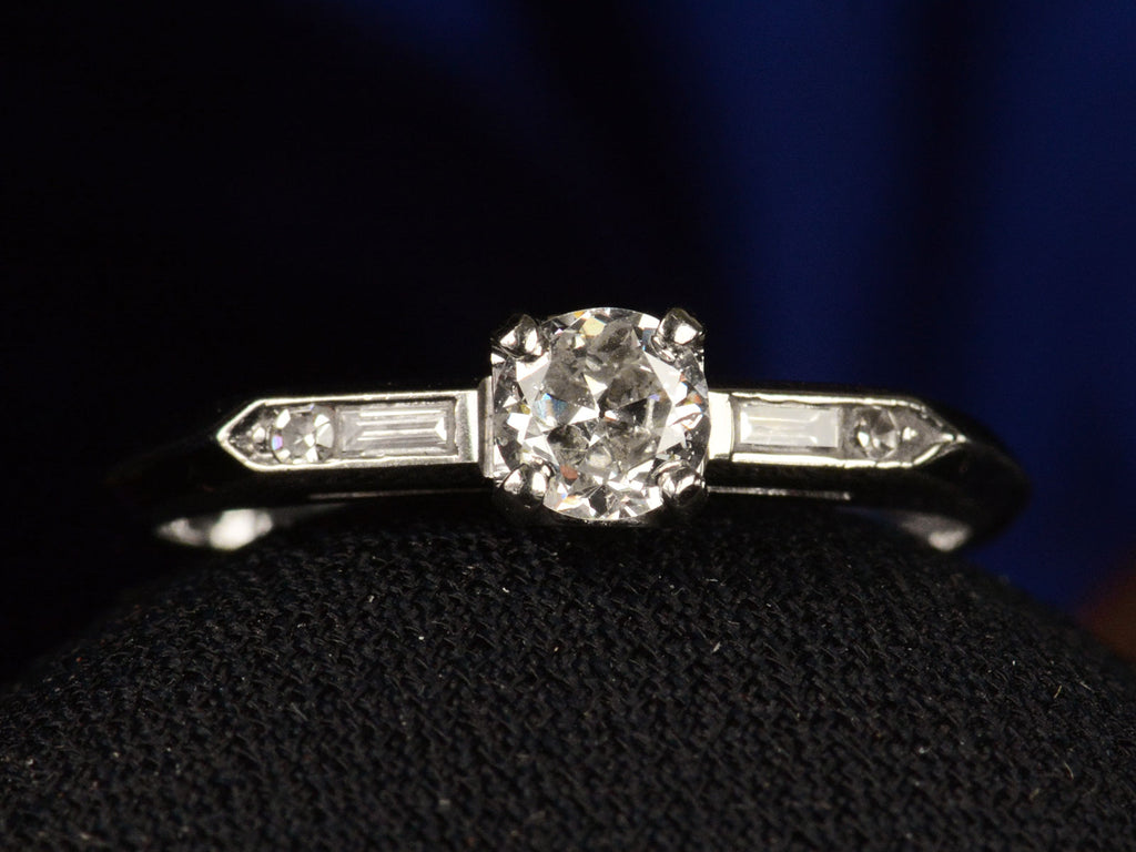 Front view of a vintage c1940 white gold engagement ring with small round diamond and rectangular baguettes on either side. Shown on dark background.