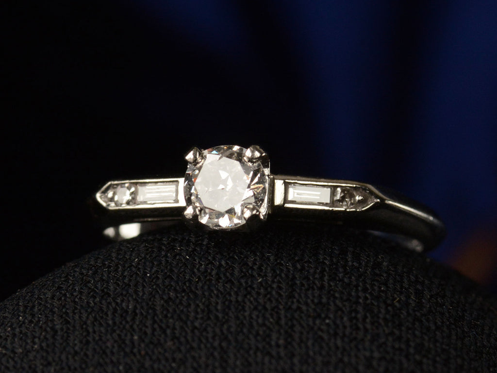 Angled front view of a vintage c1940 white gold engagement ring with small round diamond and rectangular baguettes on either side. Shown on dark background.