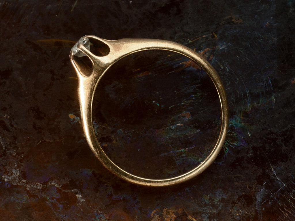 Side profile view of an antique Victorian solitaire engagement ring with a round diamond set in a six pronged rosy-yellow gold belcher mounting. Shown on dark background.