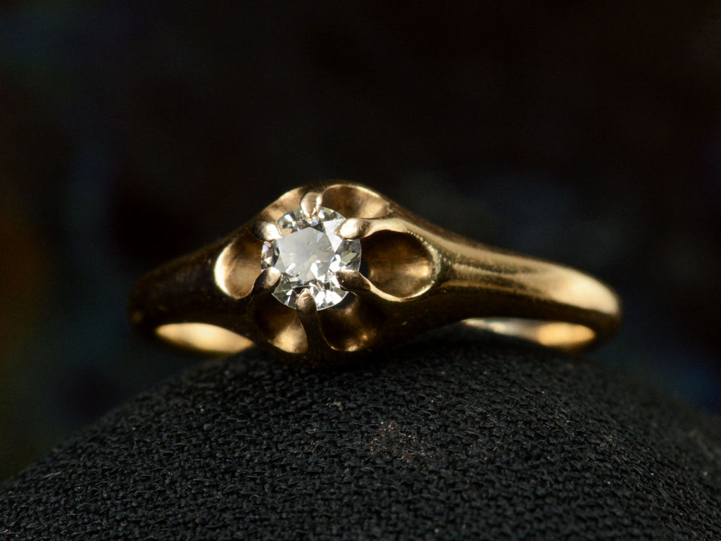 Right angled front view of an antique Victorian solitaire engagement ring with a round diamond set in a six pronged rosy-yellow gold belcher mounting. Shown on dark background.