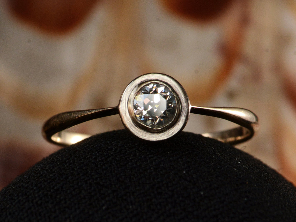 Front profile of an antique Edwardian era engagement ring with a round diamond in a simple silver topped rosy-yellow gold mounting, shown on dark background.