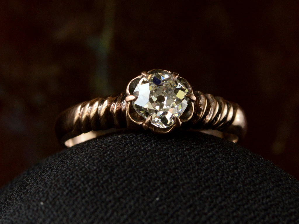 Detail of diamond in a Victorian solitaire ring with a square old mine cut diamond set in a wide striped rose gold setting, shown on dark background.