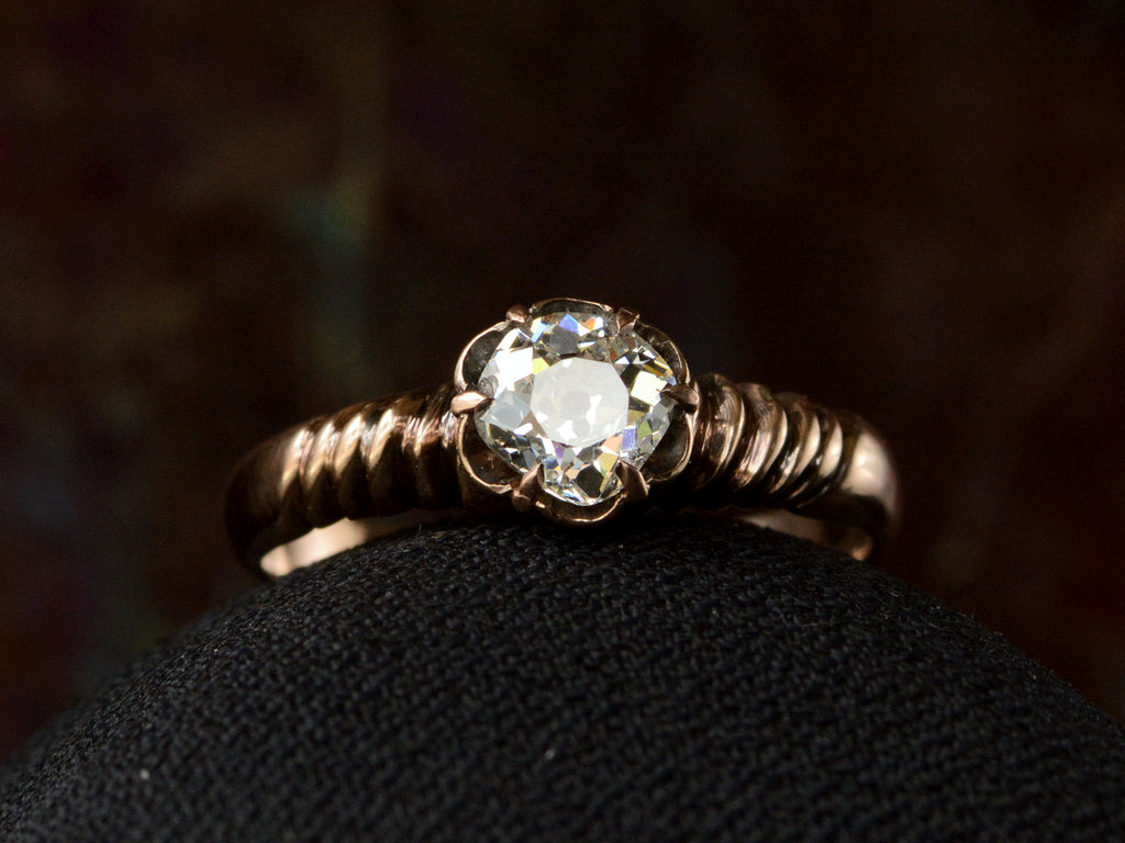 A Victorian solitaire ring with a square old mine cut diamond set in a wide striped rose gold setting, shown on white background.