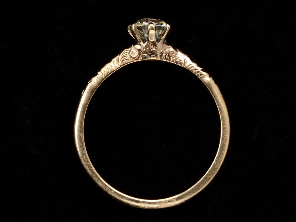 Side profile of Victorian solitaire with a slightly oval shaped diamond set in a pattered slender rose gold mounting, shown on dark background.