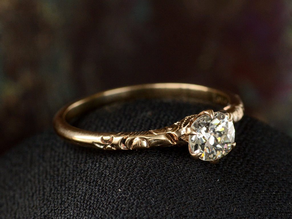 Elevated angled view of Victorian solitaire with a slightly oval shaped diamond set in a pattered slender rose gold mounting, shown on dark background.