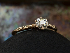 thumbnail of Left angled view of Victorian solitaire with a slightly oval shaped diamond set in a pattered slender rose gold mounting, shown on dark background.