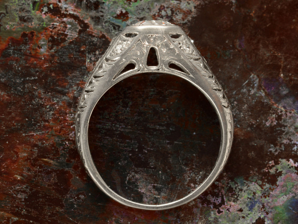 Side profile of c1920 Art Deco engagement ring with a round white diamond in a wide filigreed platinum mounting (shown on textured dark background)