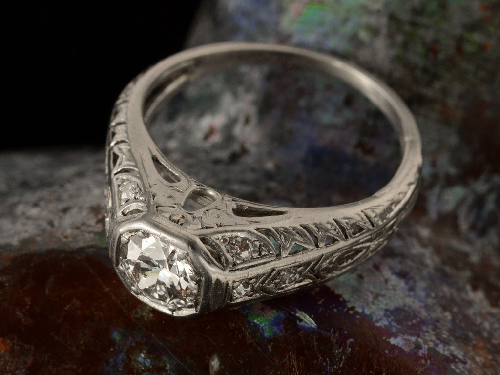 Angled top view of a c1920 Art Deco engagement ring with a round white diamond in a wide filigreed platinum mounting (shown on dark textured background)