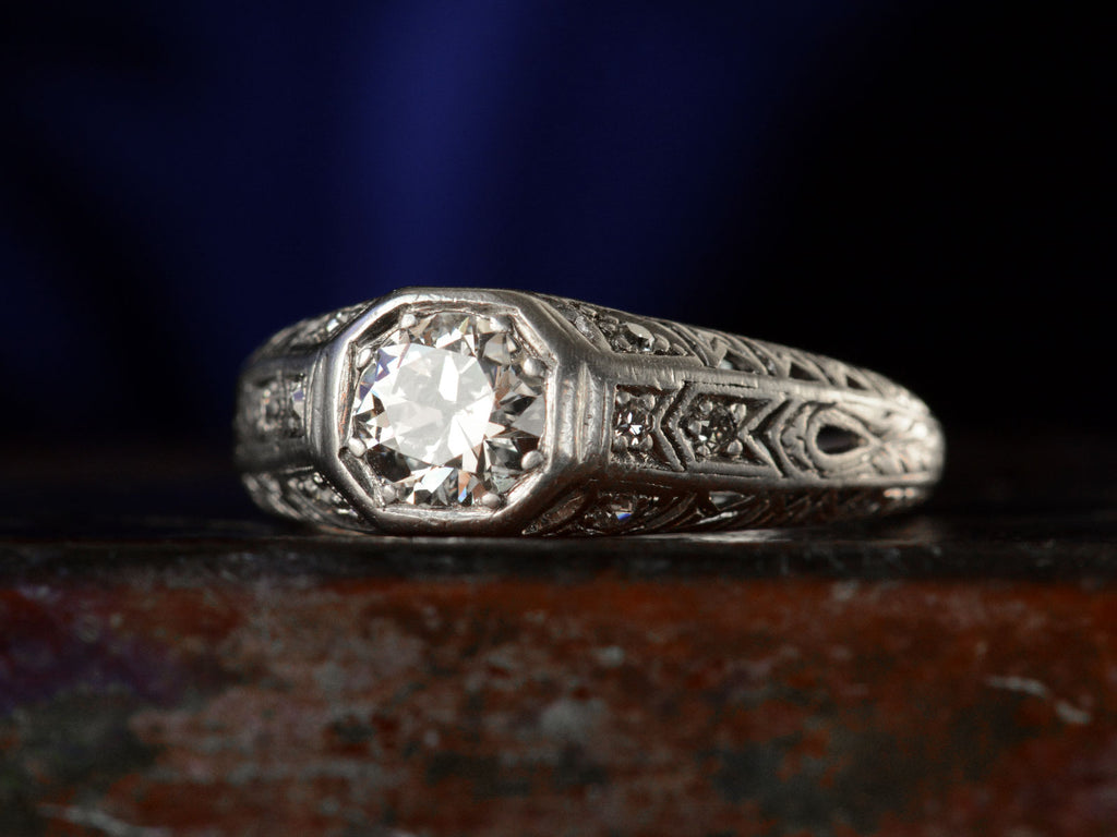Right angled view of a c1920 Art Deco engagement ring with a round white diamond in a wide filigreed platinum mounting (shown on dark background)