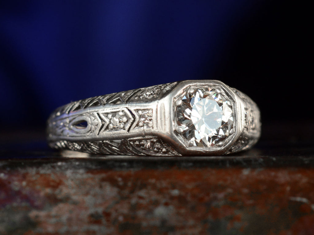 Left angled view of a c1920 Art Deco engagement ring with a round white diamond in a wide filigreed platinum mounting (shown on dark background)
