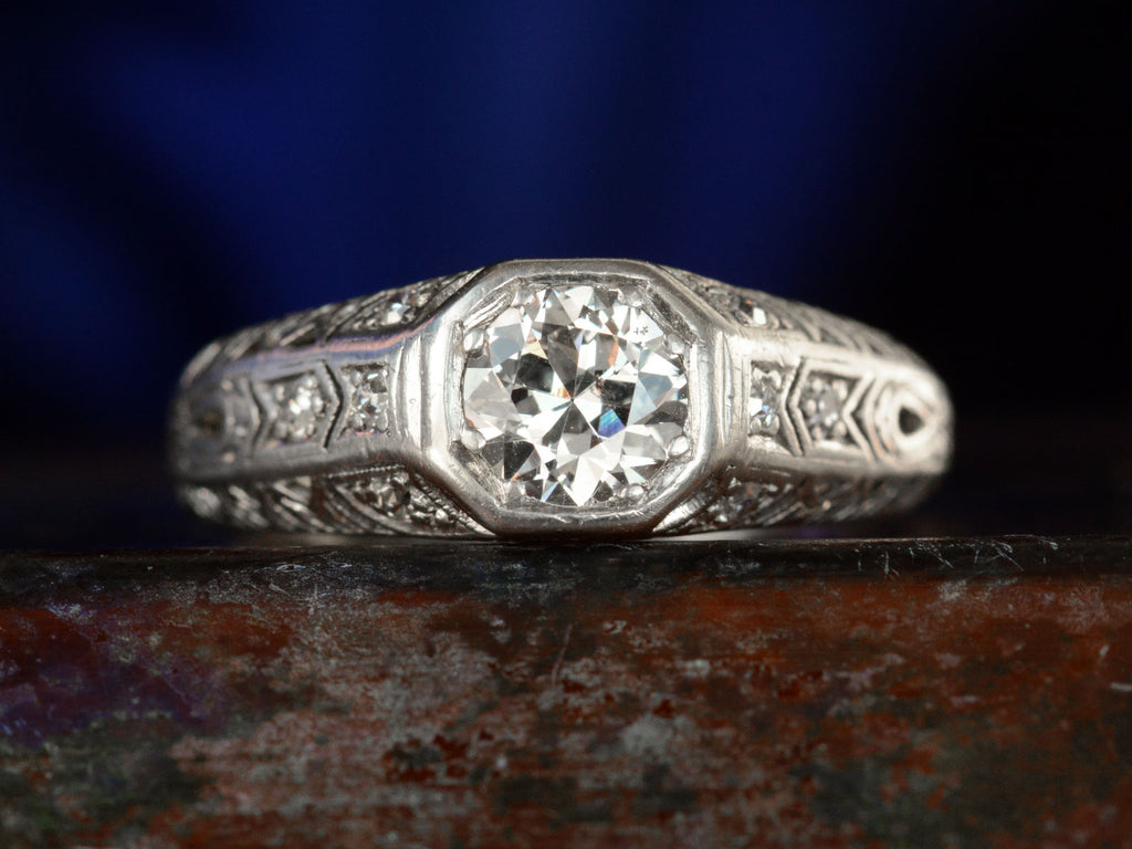 Front view of a c1920 Art Deco engagement ring with a round white diamond in a wide filigreed platinum mounting (shown on dark background)