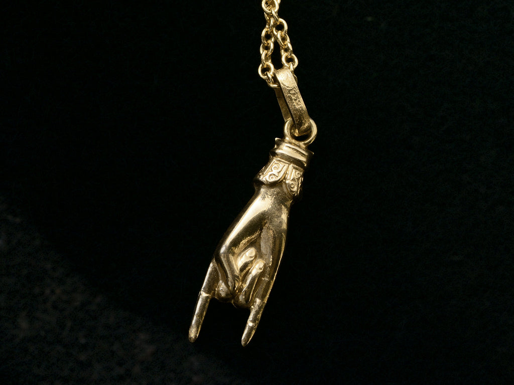 A vintage gold hand charm on a chain, gesturing the sign of the horns, shown on black background. "750" stamp visible on bale