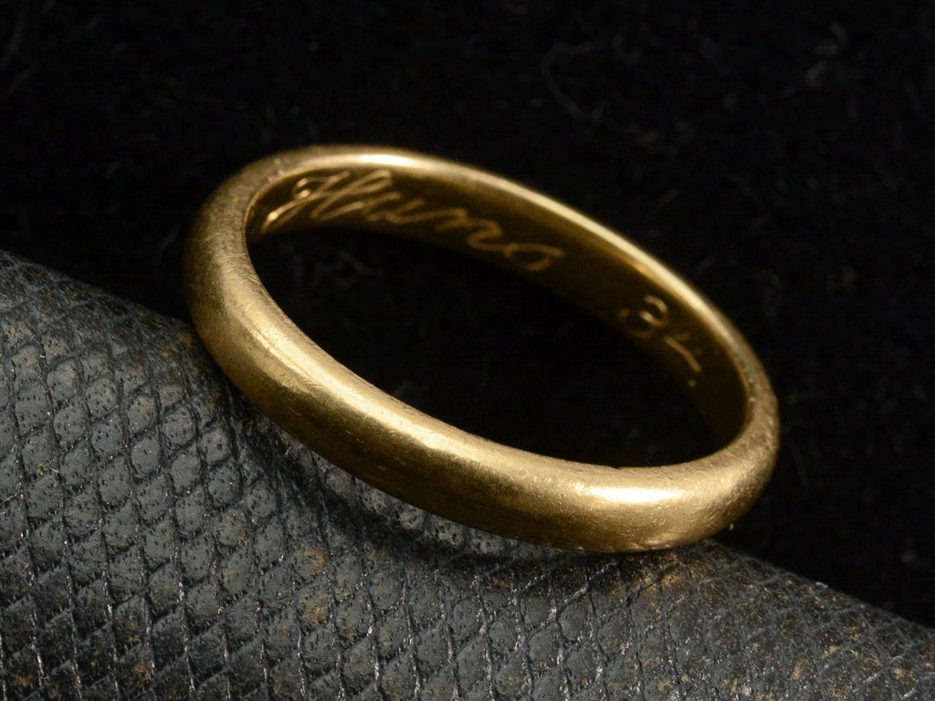 Angled view of a rounded 18K yellow gold wedding band shown on dark background. Inscription inside ring is visible: "Hans 34"