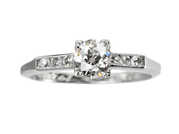 A simple Art Deco platinum engagement ring with a round old cut diamond in four prongs between 3 smaller diamonds on each side, shown on white background.
