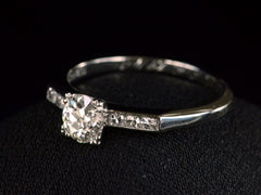 thumbnail of Right angled view of a simple Art Deco platinum engagement ring with a round old cut diamond in four prongs between 3 smaller diamonds on each side, shown on dark background.