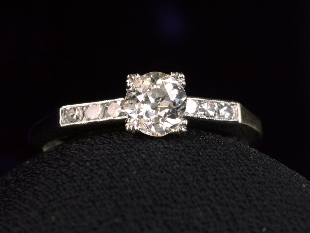 Front view of a simple Art Deco platinum engagement ring with a round old cut diamond in four prongs between 3 smaller diamonds on each side, shown on dark background.