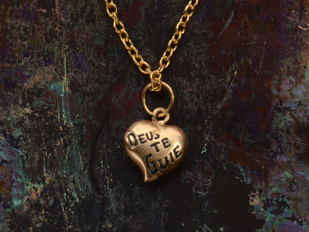 Gold heart-shaped pendant necklace with 'Deus Te Guie' text on a dark colorful background.
