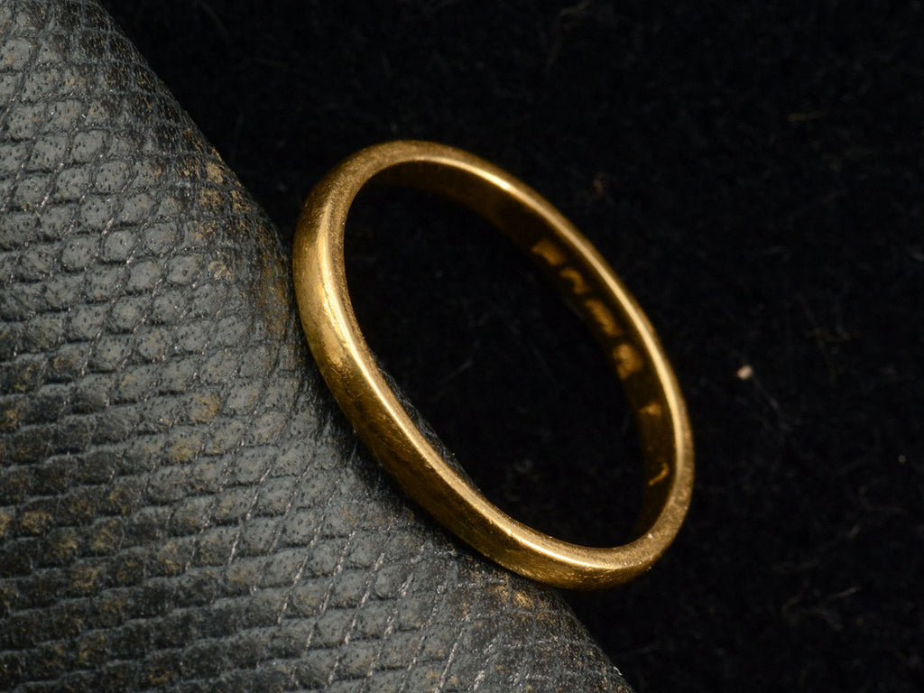 Angled view of a narrow slightly rounded 22K yellow gold band, shown on dark background with indecipherable stamps visible inside band.