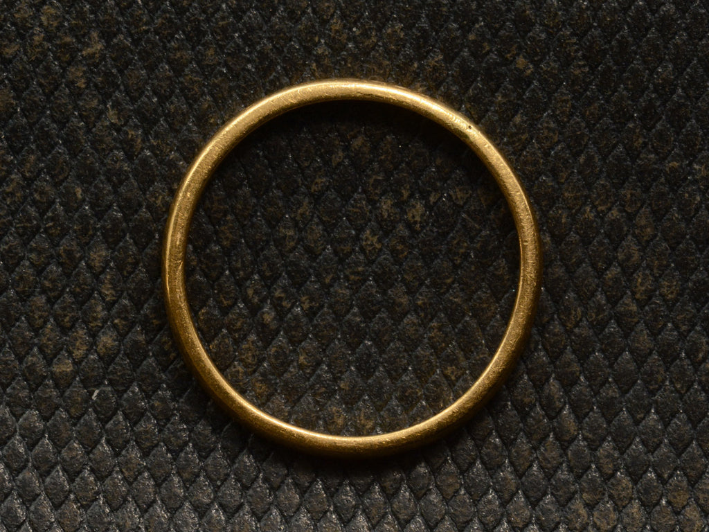 Side profile of a narrow slightly rounded 22K yellow gold band, shown on dark textured background.