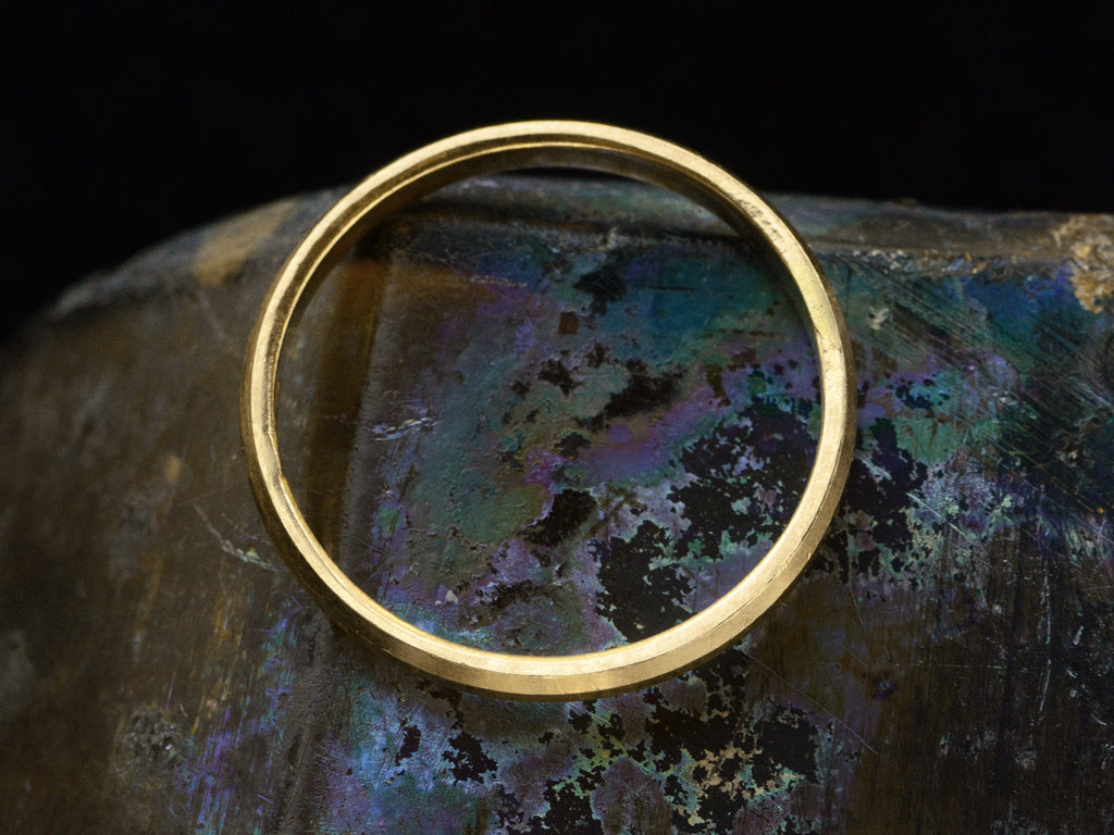Side profile of narrow 18K yellow gold band with beveled edges, shown on dark iridescent background.