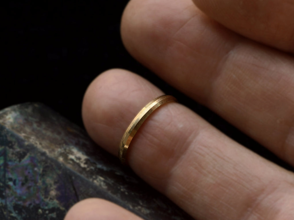 A narrow 18K yellow gold band with beveled edges, shown on finger.