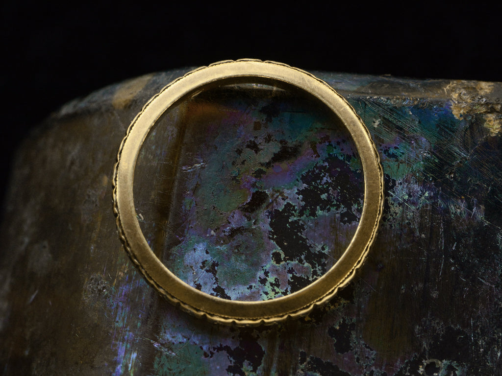 Side profile of a c1920 thin yellow gold Art Deco wedding band with patterning and white gold accents, shown against dark iridescent background.