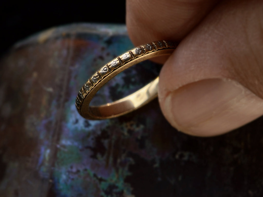 Fingers holding a c1920 thin yellow gold Art Deco wedding band with patterning and white gold accents, shown against dark textured background.