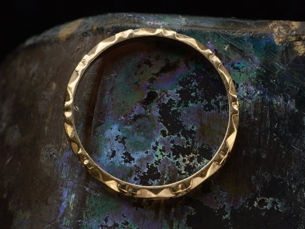 Side profile of a narrow vintage rosy-yellow wedding band with a pattern of flowers and facets, shown on a dark iridescent surface.