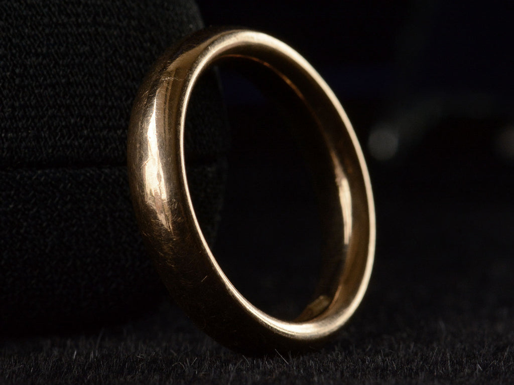 Angled view of rounded 4.0mm wide antique rosy-yellow gold wedding band, shown on black background.