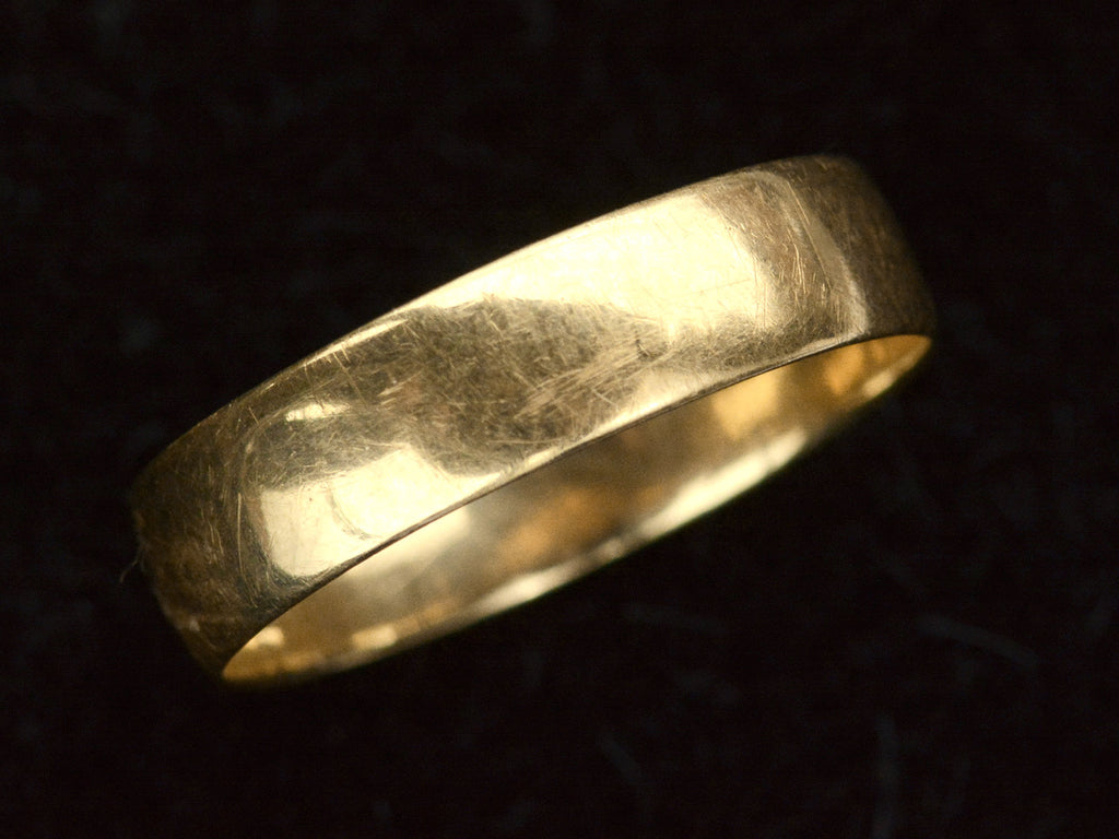 Angled view of wide flat 18K yellow gold antique wedding band, on dark background.