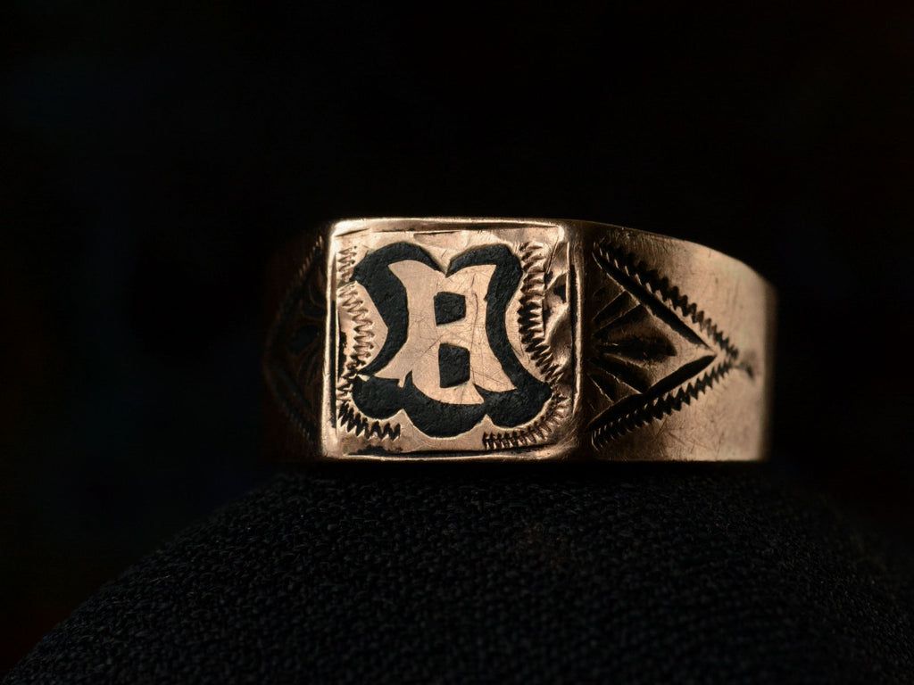 Angled front view of antique c1890 Victorian signet ring in rose gold with black enamel details and the letter "B" on the face. Shown on dark background.
