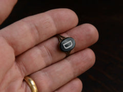 thumbnail of c1890 Victorian signet ring with oval black plaque and white letter "D". Set in brass and shown on hand for scale.