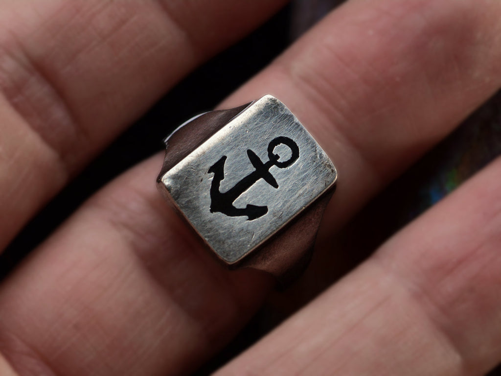 A sterling silver rectangular signet ring with the image of an anchor enameled in black, shown on finger.