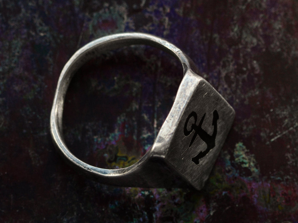 Side profile view of a sterling silver rectangular signet ring with the image of an anchor enameled in black, shown on dark background.