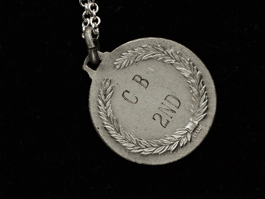 Reverse side detail of a vintage white metal archery medal inscribed "C B 2ND" on the back with an "ITALY" mark also showing. Shown on black background.