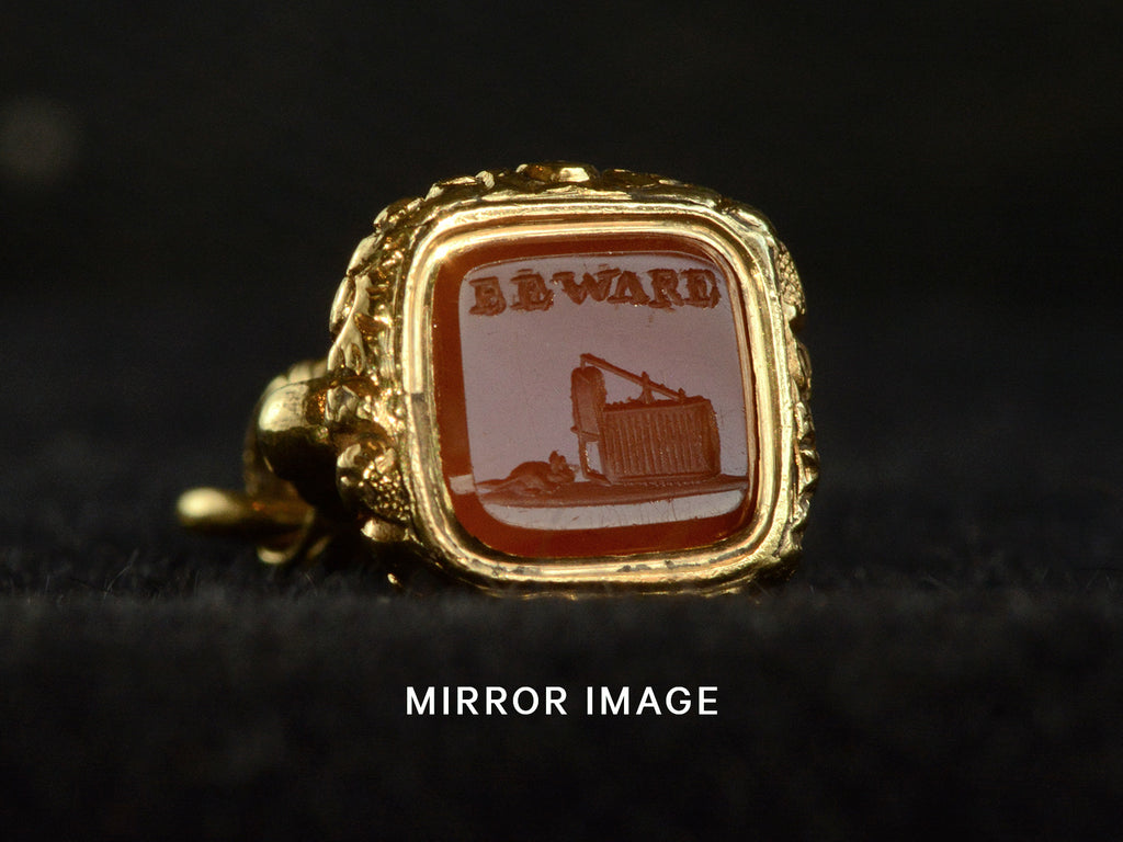 Angled view of Georgian-era gold fob with a carnelian intaglio carved with the word "BEWARE" in and the image of a mousetrap and mouse, shown on dark background. Captioned with "MIRROR IMAGE" indicating that the image has be mirrored to show how the image would look impressed in wax.