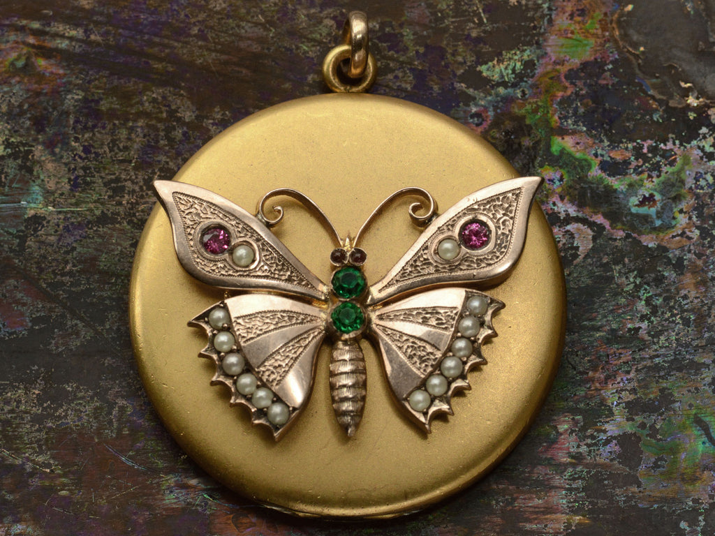 A round Victorian gilt locket with a rose gold butterfly mounted on the front, detailed with pearls and red and green paste. Shown on dark background.