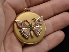 thumbnail of A round Victorian gilt locket with a rose gold butterfly mounted on the front, detailed with pearls and red and green paste. Shown on hand