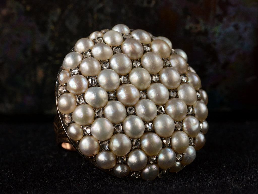 Angled left view of Victorian ring with a large luster pearls and rose cut diamonds, set in rosy-yellow gold, shown on dark background.