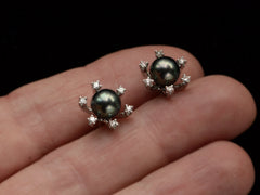 thumbnail of A  pair of vintage starburst stud earrings featuring a dark green-gray cultured pearls in a white gold prong setting, surrounded by six small clear diamonds on a hand.
