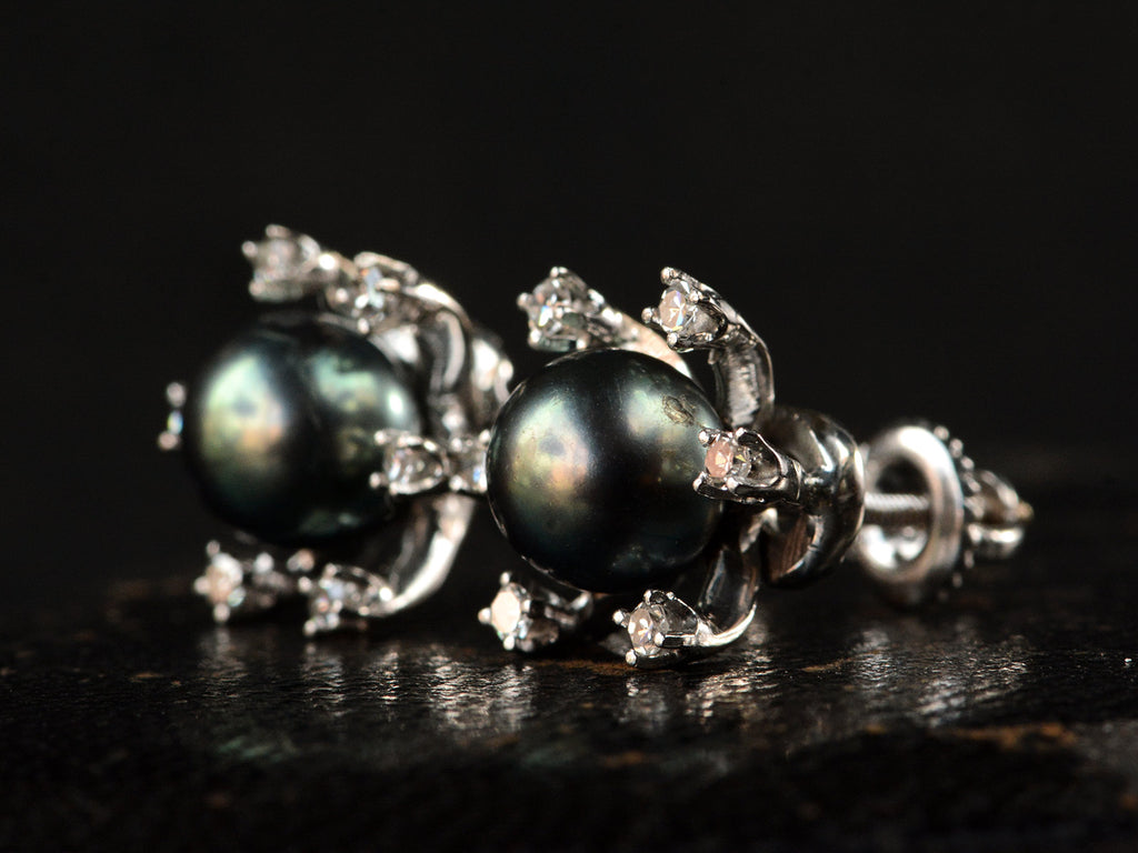 Angled view of a pair of vintage starburst stud earrings featuring a dark green-gray cultured pearls in a white gold prong setting, surrounded by six small clear diamonds on a dark background.