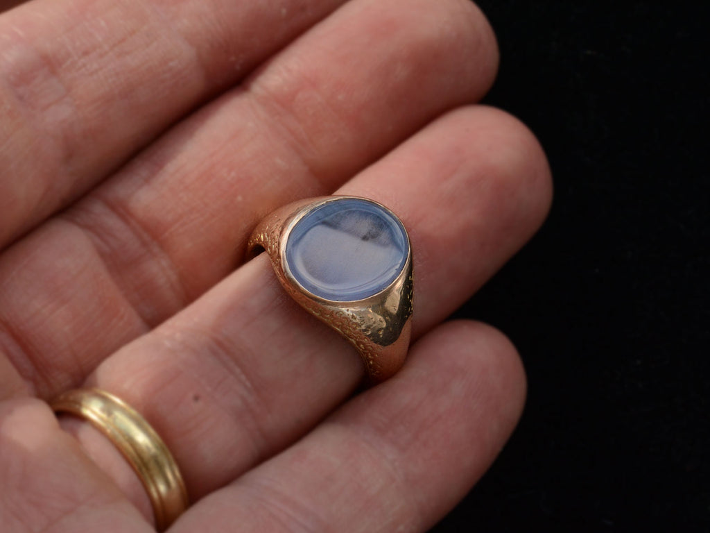 Angled view of antique gold signet ring with oval blue chalcedony face, on finger.