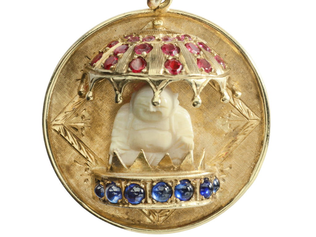 A round  yellow gold vintage pendant with a carved bone Buddha figure beneath a parasol of rubies and resting on a sapphire lined dais. Shown on white background.