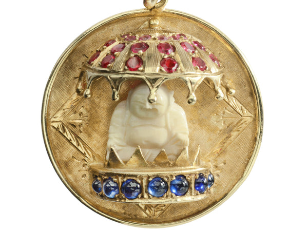 A round  yellow gold vintage pendant with a carved bone Buddha figure beneath a parasol of rubies and resting on a sapphire lined dais. Shown on white background.