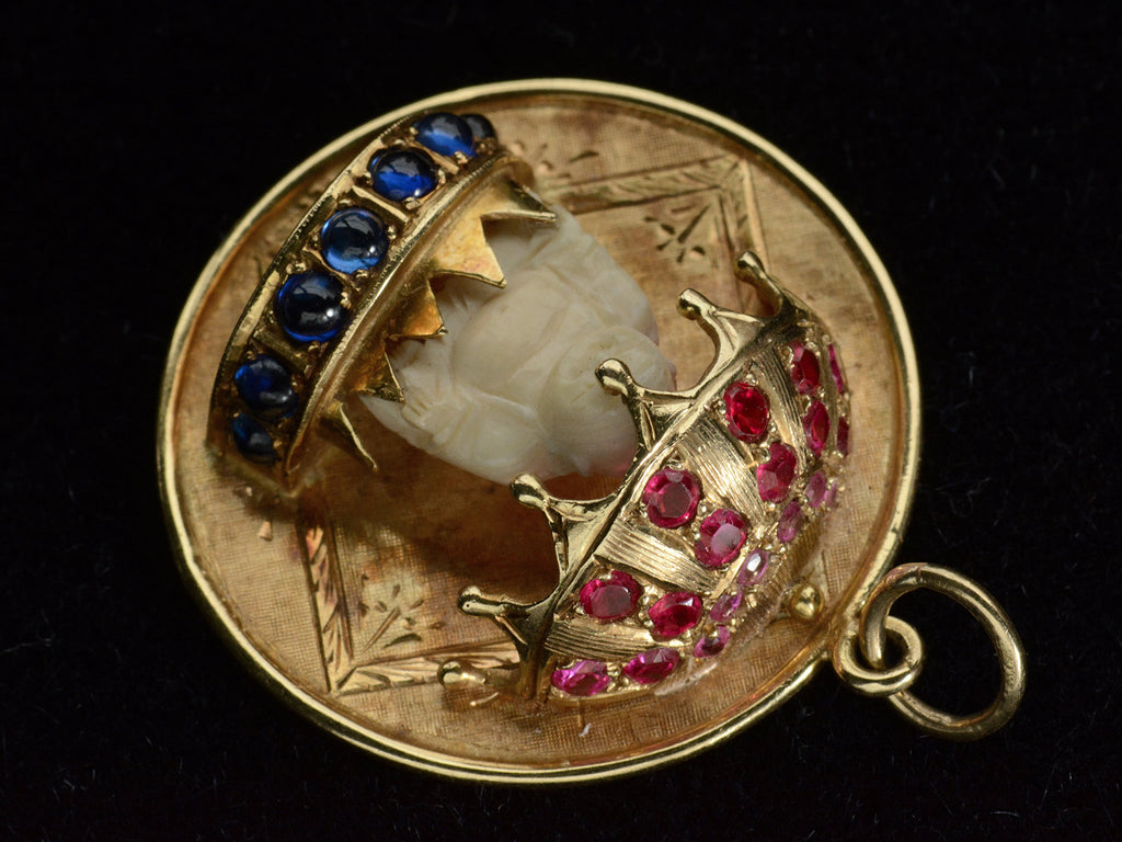 Reverse angled view of a round  yellow gold vintage pendant with a carved bone Buddha figure beneath a parasol of rubies and resting on a sapphire lined dais. Shown on black background.
