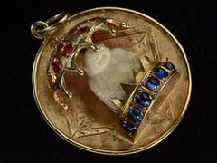thumbnail of Angled view of a round  yellow gold vintage pendant with a carved bone Buddha figure beneath a parasol of rubies and resting on a sapphire lined dais. Shown on black background.